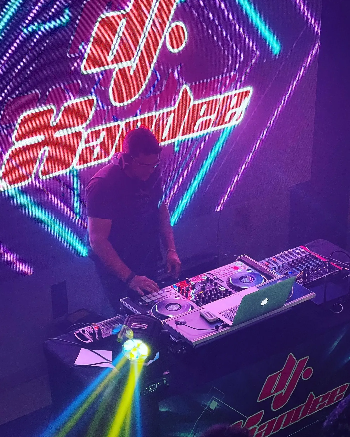 DJ Xandee tocando na cabine em frente à logo em neon vermelho e roxo